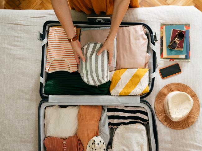 Shot from above of an anonymous woman packing things in her suitcase on the bed. She is holding and putting a grey and white stripped cosmetics bag in. There is her mobile phone, books and sunglasses on the bed.Escape 7 May 2023Doc HolidayPhoto - iStock