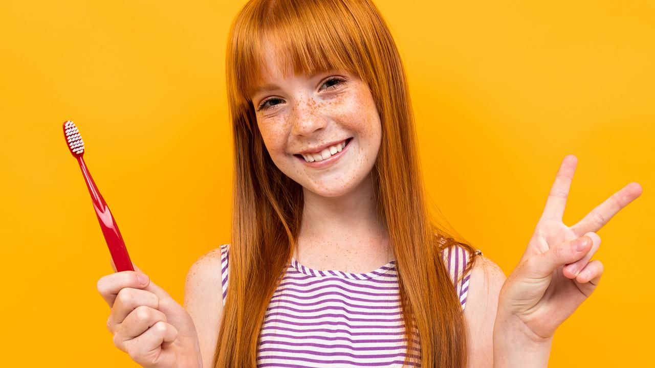 red-haired girl holds a toothbrush in hands over yellow background.