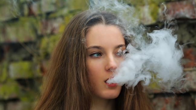 Vape teenager. Young pretty white girl in white cardigan smoking an electronic cigarette opposite destroyed brick wall on the street in the spring. Bad habit that is harmful to health.