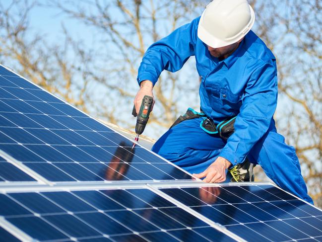 Male engineer in blue suit and protective helmet installing photovoltaic panel system using screwdriver. Professional electrician mounting solar module on roof. Alternative energy ecological concept.
