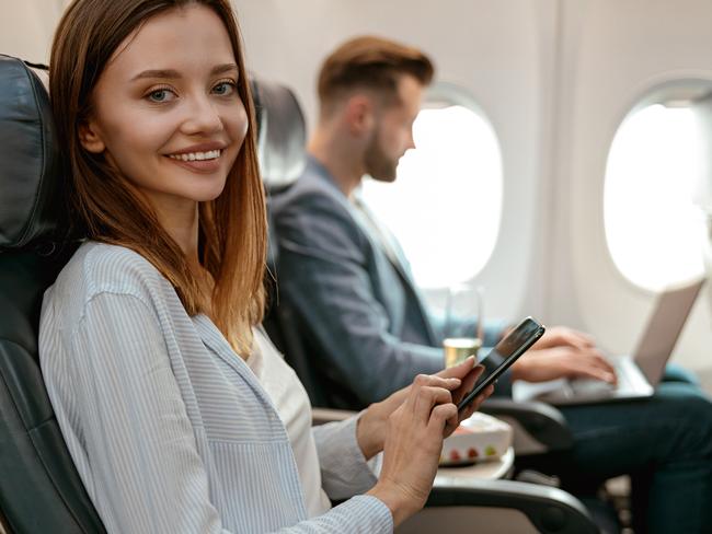 Cheerful female traveler holding smartphone and smiling while sitting in passenger chair in plane