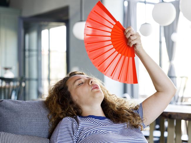 Young woman sit on couch at home waving with paper fan cooling down, sweating woman relax on sofa in living room