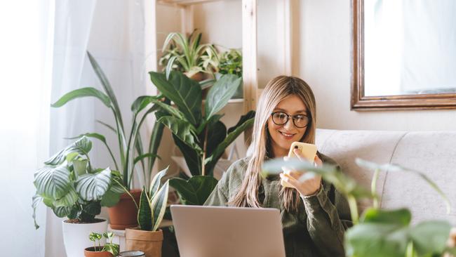 Freelance woman in glasses with mobile phone typing at laptop and working from home office. Happy girl sitting on couch in living room with plants. Distance learning online education and work.