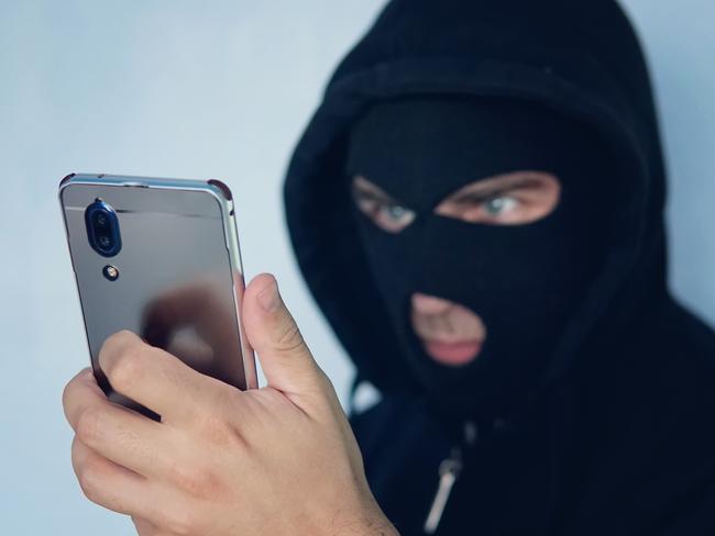 Young adult in black clothes with hidden face looks at smartphone screen. Ill-intended fraudster uses mobile. Fraudster calls. Mobile racket. Hacker hijacks by phone. Cellphone account fraud. Scam