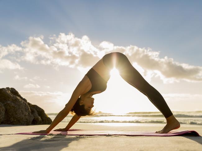 Side view of female athlete doing yoga in downward facing dog position on shore. Sunlight is streaming through young woman exercising at beach during sunset. She is in sportswear.