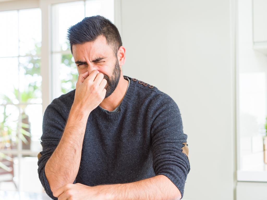 Handsome hispanic man wearing casual sweater at home smelling something stinky and disgusting, intolerable smell, holding breath with fingers on nose. Bad smells concept.
