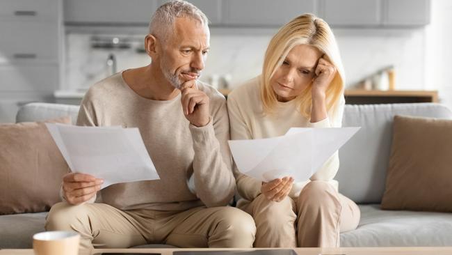 Mature couple looking at documents with concern, sitting on a couch with a tablet and coffee cup; stressed investors bills middle-aged generic