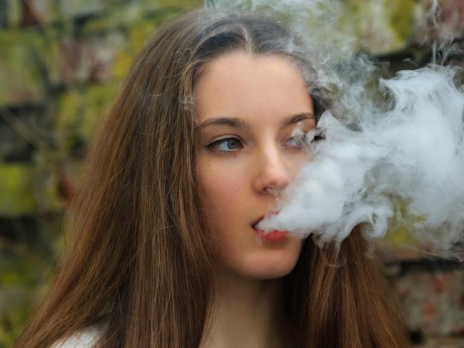 Vape teenager. Young pretty white girl in white cardigan smoking an electronic cigarette opposite destroyed brick wall on the street in the spring. Bad habit that is harmful to health.