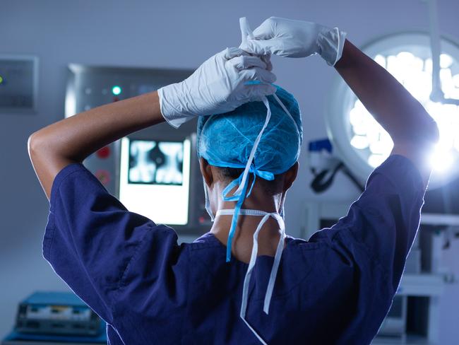 Rear view of young African-american female surgeon putting her surgical mask on in operating room at hospital. Shot in real medical hospital with doctors nurses and surgeons in authentic setting