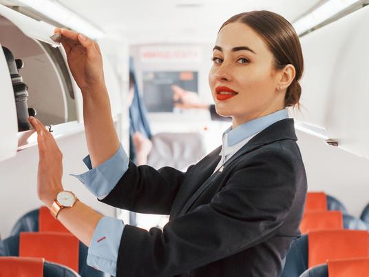 Young stewardess that is in formal black clothes is standing indoors in the plane.