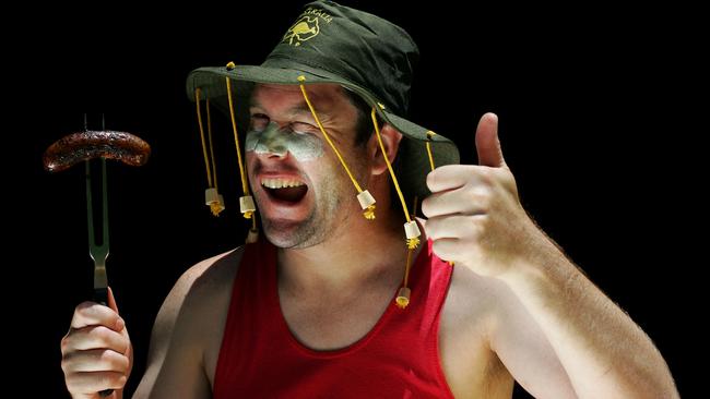 An Aussie man in a cork hat winks at the camera holding up a sausage on a fork in one hand and giving the thumbs up gesture with the other