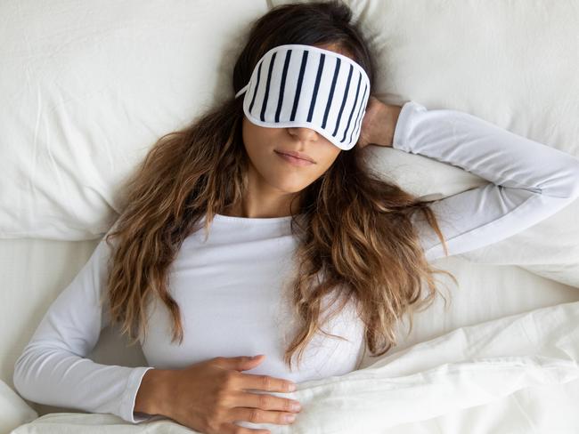 Calm peaceful mixed race young lady wearing sleeping mask, enjoying night dream, napping in comfortable bed at home. Top above view serene biracial millennial woman relaxing on pillow under blanket.
