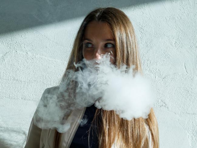 Vape teenager. Young cute girl in  casual clothes smokes an electronic cigarette near the wall outdoors in summer day. Bad habit that is harmful to health. Vaping activity.