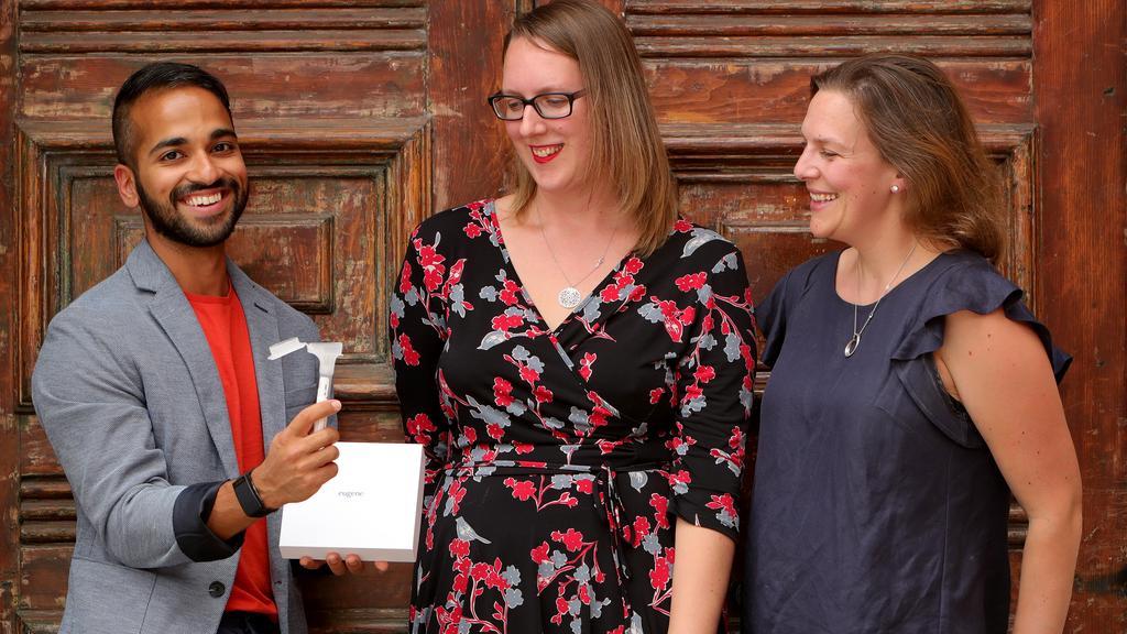 Three people pose for a picture. The man on the left is holding an item, whereas the two others are looking at it. All are smiling.