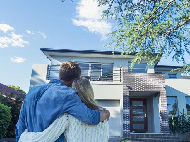 Couple standing in front of their new home. They are both wearing casual clothes and embracing. Rear view from behind them. The house is contemporary with a brick facade, driveway, balcony and a green lawn. The front door is also visible. Copy space