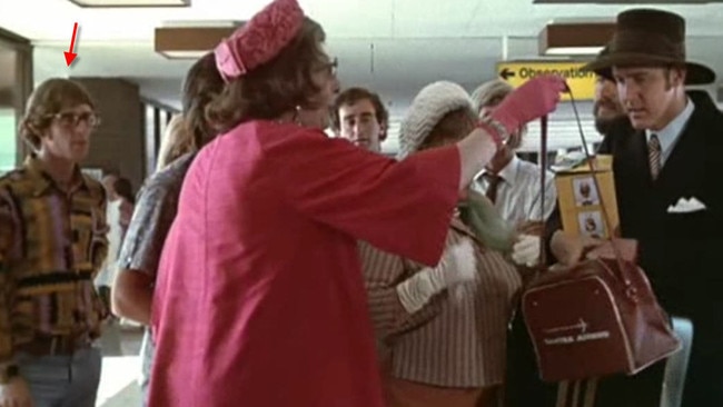 Ross Fitzgerald (red arrow) looks on during his 90-second cameo as Barry Humphries in character as Dame Edna Everage hands Barry Crocker a flight bag in a scene from The Adventures of Barry McKenzie.