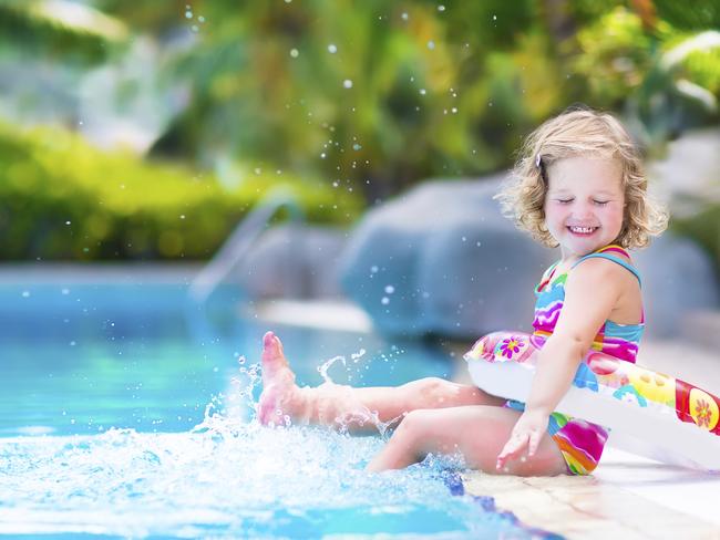 Adorable little girl with curly hair wearing a colorful swimming suit playing with water splashes at beautiful pool in a tropical resort having fun during family summer vacation