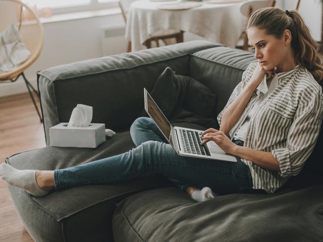 Early symptoms of disease. Side on full length portrait of tired sick lady watching medical program on laptop while sitting on sofa with nasal paper wipes Picture: Istock