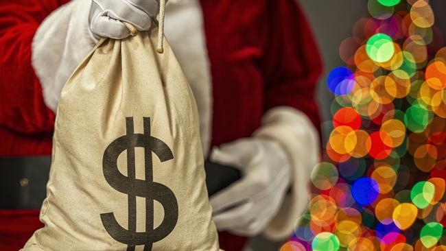 Santa Claus holding a large canvas money bag with black dollar sign symbol and in the background the bokeh of multi-colored Christmas tree lights