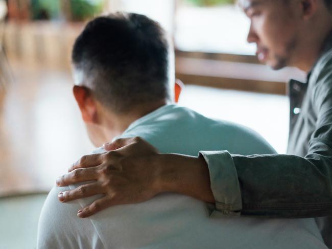 Rear view of son and elderly father sitting together at home. Son caring for his father, putting hand on his shoulder, comforting and consoling him. Family love, bonding, care and confidence