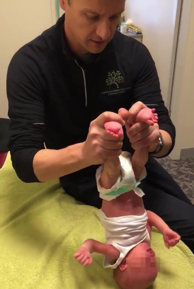 Chiropractor Dr Andrew Arnold briefly raises the infant by the ankles while checking its neck, before asking another person off camera to help cradle its neck as he lays it back on the examination bed.