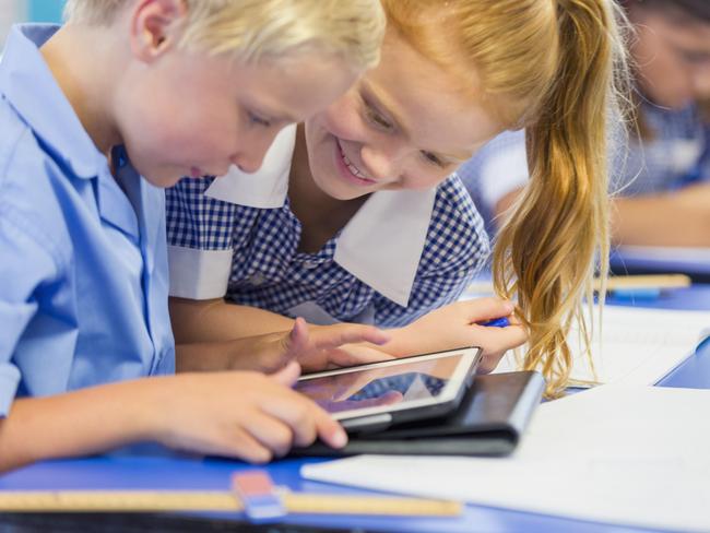 Boy and girl students sharing a digital tablet. They are in a classroom wearing school uniforms. Both are excited, and one is smiling. Ethnic students in the background.