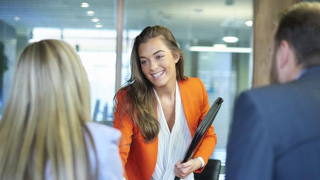 a young graduate walks into an interview room full of confidence and positivity energy . She is holding her cv and smiling at the interview panel before her. She is wearing blue trousers with an orange suit jacket , as she opens the door and strides in and shakes hands with a woman on the panel . in the foreground we can see the back of two of the panel , a man and a woman.