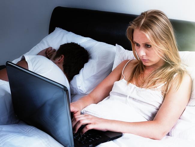 A handsome young couple lie in bed; she's intent on her laptop, either keeping up with social media, working late, or battling insomnia; he's fast asleep next to her. Istock