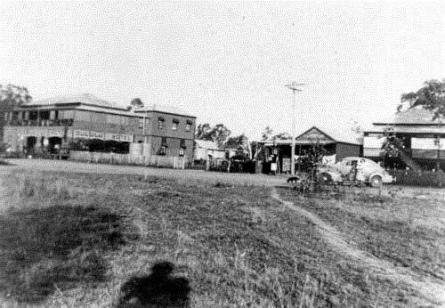 Exterior view of the Dululu Hotel, Dululu. The fiture of a friendly man wearing a stockman's hat is said to haunt the Banana Shire pub.Caption: Behind the hotel building can be seen small two residential buildings, both of which have corrugated galvanised iron tanks. To the right of the hotel is a commercial building that has a petrol pump out the front.