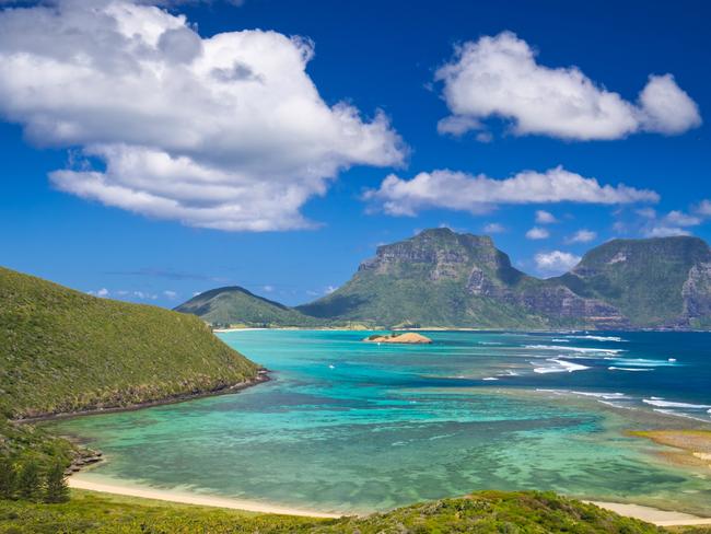 View south over the stunning turquoise lagoon to the peaks of Mts Lidgbird and Gower. Taken from Mt Eliza on Lord Howe Island, Australia. Fluffy clouds stand out from the deep blue sky overhead. This tiny island is a delightful tourist destination.Escape 11 August 2024Why I TravelPhoto - iStock