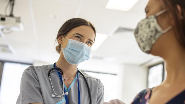 Shot focused on a kind looking nurse wearing scrubs, a stethoscope, white rubber gloves and a protective face mask. She is smiling at the nervous patient with her eyes. The nurse is prepping the patients arm before she injects her with the COVID-19 vaccine.