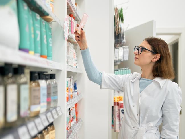 Woman in white robe and glasses taking bottle of medical cream from shelf while working in modern pharmacy.
