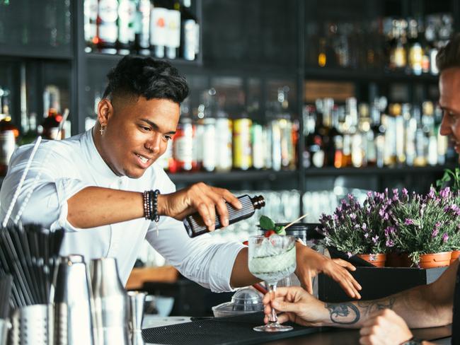 Mixed race male expert bartender is serving some tonic for a cocktail at the bar counter while a smiling waiter is holding the cocktail glass