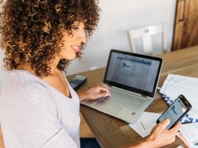 A woman sits at her dining room table with laptop computer and financial reports doing her monthly budget. She is smiling at the ease of use as she works on her smart phone banking app to do monthly finances, pay taxes and save money for the future. She is also filing her tax return. Picture: iStock.