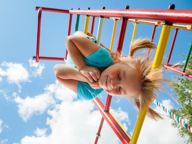 children playing  Low angle view of cute blond girl wearing blue tshirt hanging from a monkey bars. Girl is smiling with her eyes closed. The climbing frame is painted in red yellow green colors and located in the courtyard of a house. Blue summer sky with clouds and tree leaves are seen in the background.