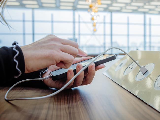 Close-up of a woman with blond hair, wearing a black pullover, using her smartphone while charging it via a white USB cable on a public charging station installed on a wooden table inside an airport terminal, window of the terminal in the background, focus on forefront, horizontal  Picture: istock