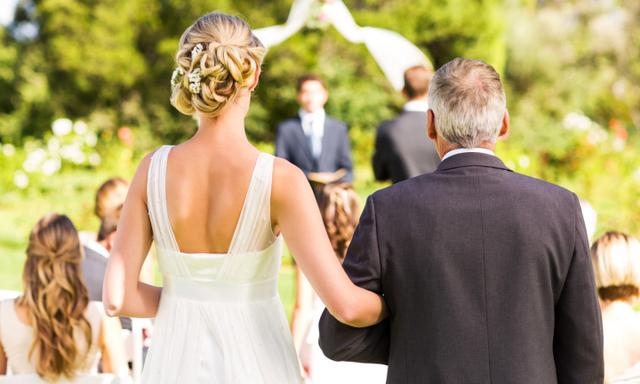 Rear view of bride and father walking down the aisle with people in background during outdoor wedding. Horizontal shot.
