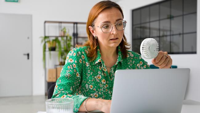 Tired mature businesswoman using laptop while sitting with portable electric fan at office desk