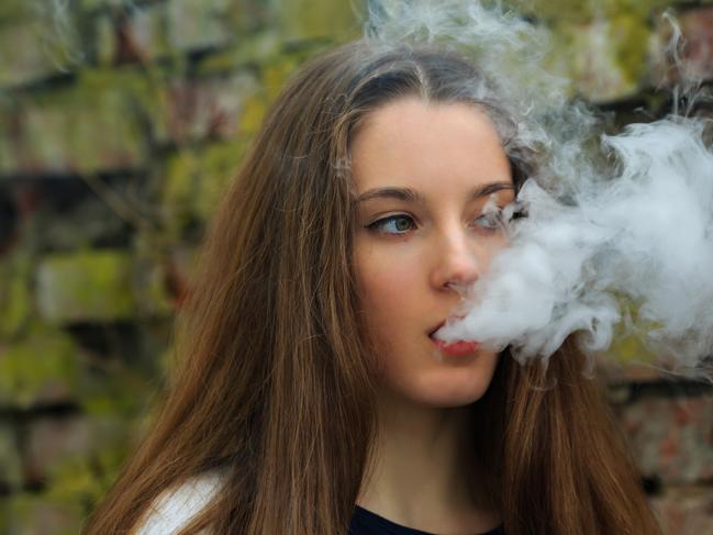 Vape teenager. Young pretty white girl in white cardigan smoking an electronic cigarette opposite destroyed brick wall on the street in the spring. Bad habit that is harmful to health.