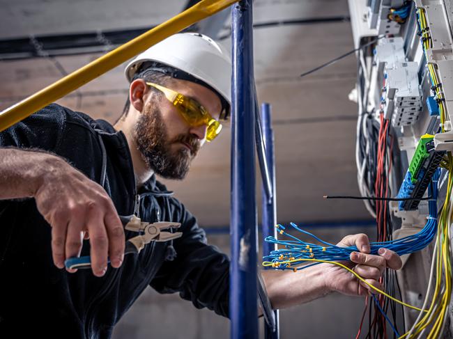 A male electrician works in a switchboard with an electrical connecting cable, connects the equipment with tools.