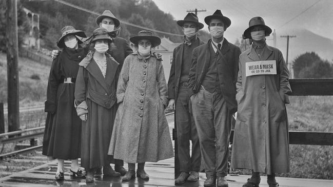 A group of people standing outdoors wearing masks over their mouths during the Spanish flu pandemic. The woman on the right wears a sign reading “wear a mask or go to jail”.