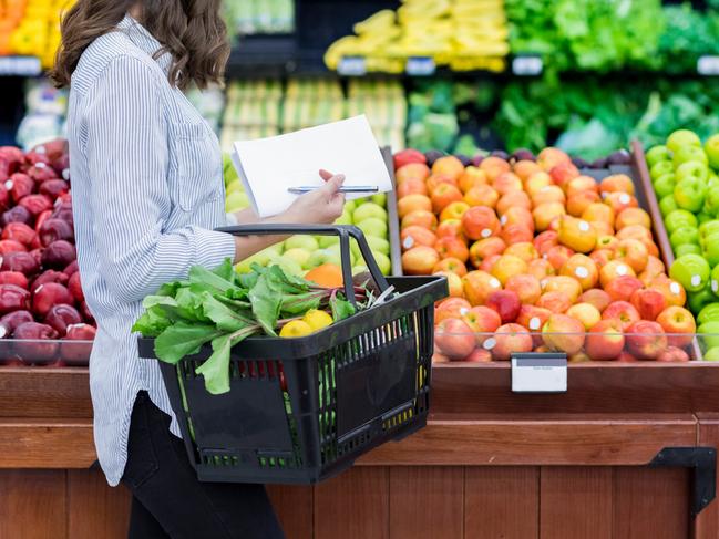 Young woman carries a shopping basket filled with fresh produce. She is shopping for fresh fruit and vegetables in a grocery store.