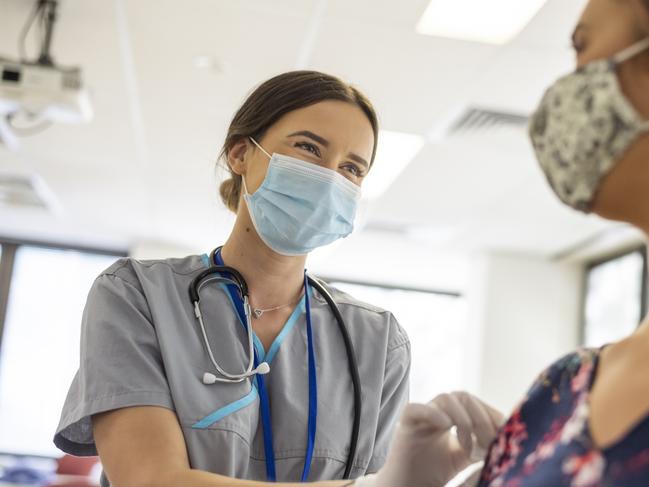 Shot focused on a kind looking nurse wearing scrubs, a stethoscope, white rubber gloves and a protective face mask. She is smiling at the nervous patient with her eyes. The nurse is prepping the patients arm before she injects her with the COVID-19 vaccine.