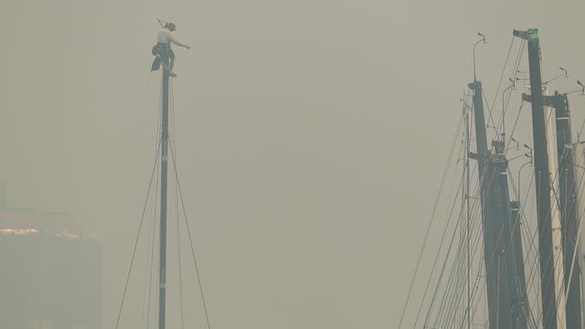 A crew member climbs the mast of a yacht docked at the Cruising Yacht Club of Australia as smoke covers Sydney Harbour. Tuesday’s Big Boat Challenge on the harbour has been abandoned. Picture: Brett Costello