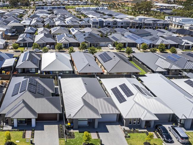 Low aerial close view new dense rural housing development, mostly grey roofing, some green landscaping, young trees. Rows of houses of various architectural styles with shades of grey roofing in rural urban sprawl, some still under construction.  Tightly-packed houses with small front and back yards and few if any trees. Some solar panels installed. Mt Barker near Adelaide, South Australia