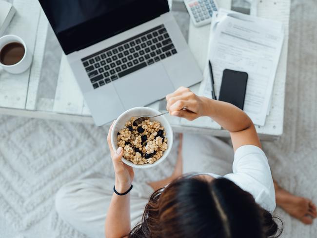 Overhead view of young Asian woman working on laptop, analysing bills and handling documents while having breakfast in the living room at home. Working from home, freelancer, home finances concept