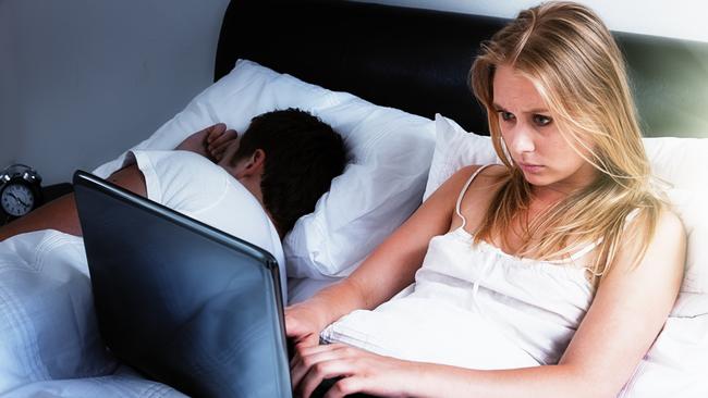 A handsome young couple lie in bed; she's intent on her laptop, either keeping up with social media, working late, or battling insomnia; he's fast asleep next to her. Istock