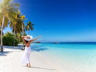 A tourist woman with white hat walks down a tropical paradise beach with palm trees, fine sand, turquoise sea and copy space in the sky