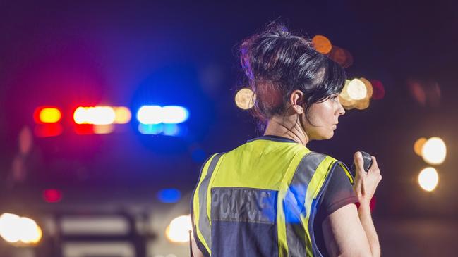 Rear view of a female police officer standing in the street at night, talking into her radio. Her patrol car is in the background with the emergency lights illuminated. She is wearing a yellow safety vest. Tablet generic