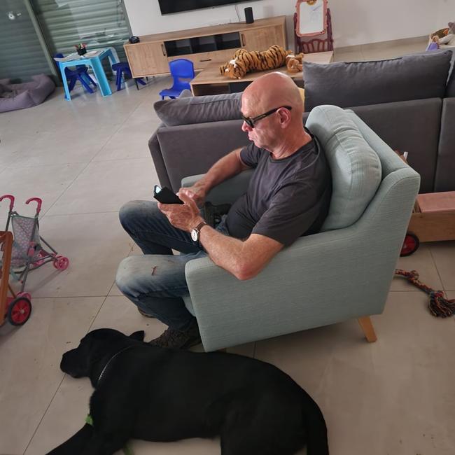 Noam Tibon holding a pistol, with the dog resting in Amir and Miri’s home in Nahal Oz, ten hours after he left his house in Tel Aviv and raced toward the Gaza border area. Bloodstains are visible on his jeans, and a bullet hole can be seen in the window at top left.